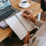 Man writing in notebook with laptop and coffee in home office setting.