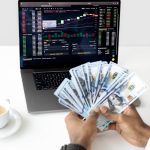Hands holding cash in front of a laptop displaying stock market data, symbolizing financial growth and investment.